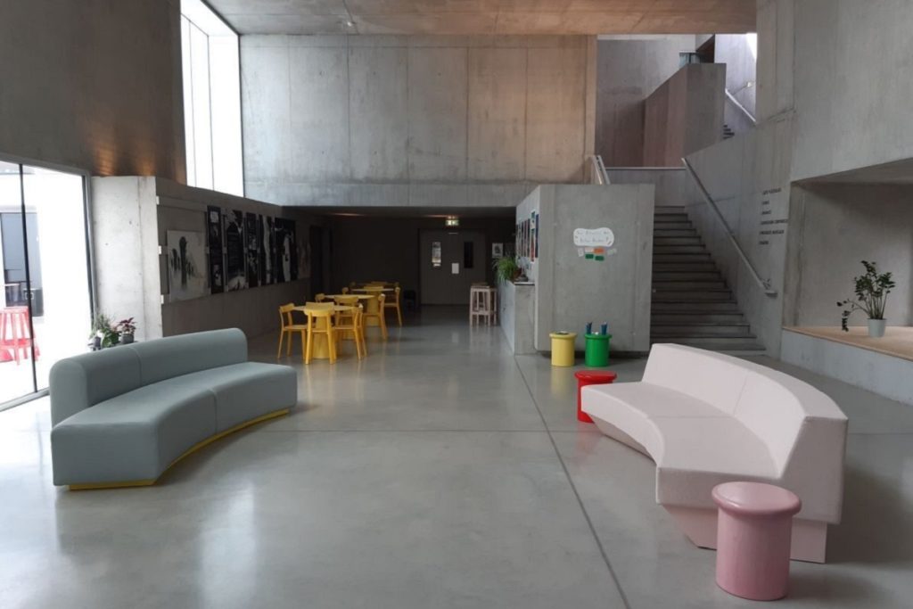 Hall d'entrée Agora, espace commun à la MJC et à la médiathèque, avec canapés courbes, tables et escalier menant à l'étage.