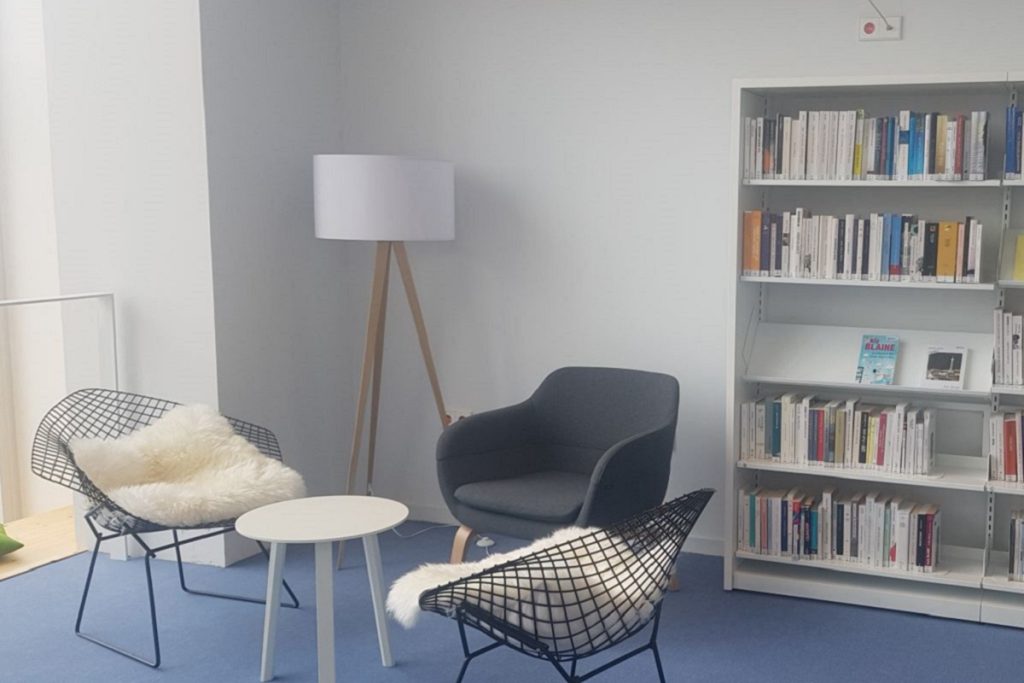 Coin lecture de l'espace adulte avec deux fauteuils filaires garnis de peaux de mouton, un fauteuil gris et un lampadaire, face à une étagère de livres.