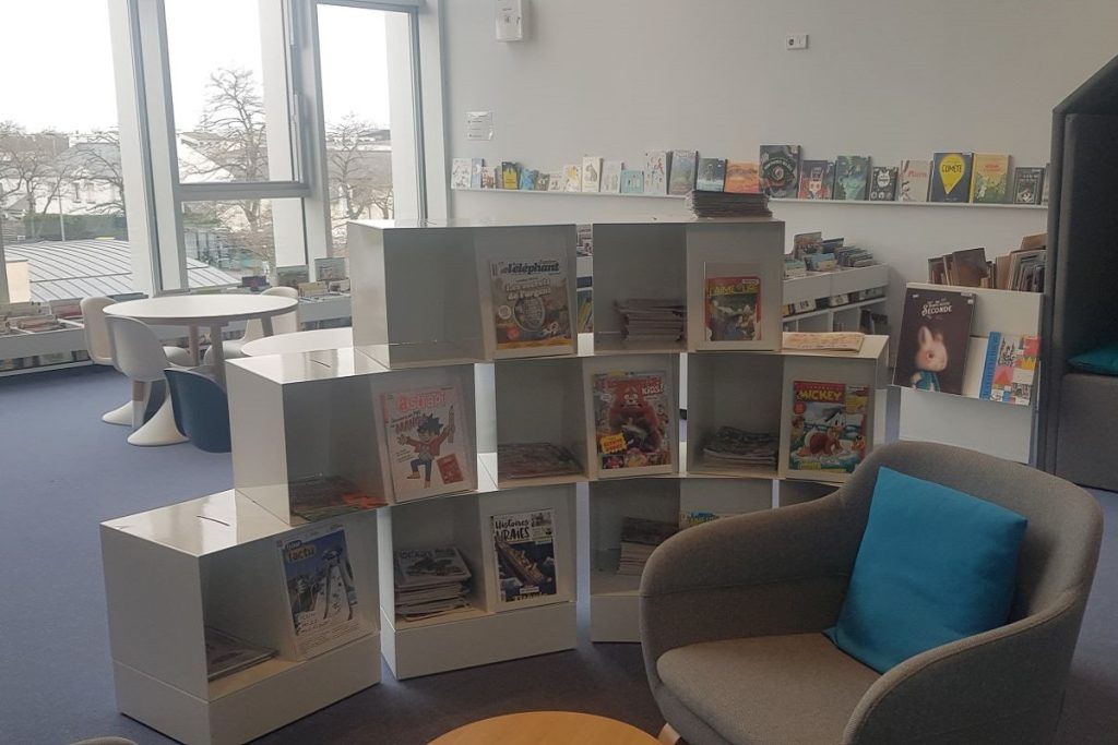 Espace jeunesse avec meubles bas empilés présentant des magazines et bandes dessinées, et un fauteuil à coussin bleu.
