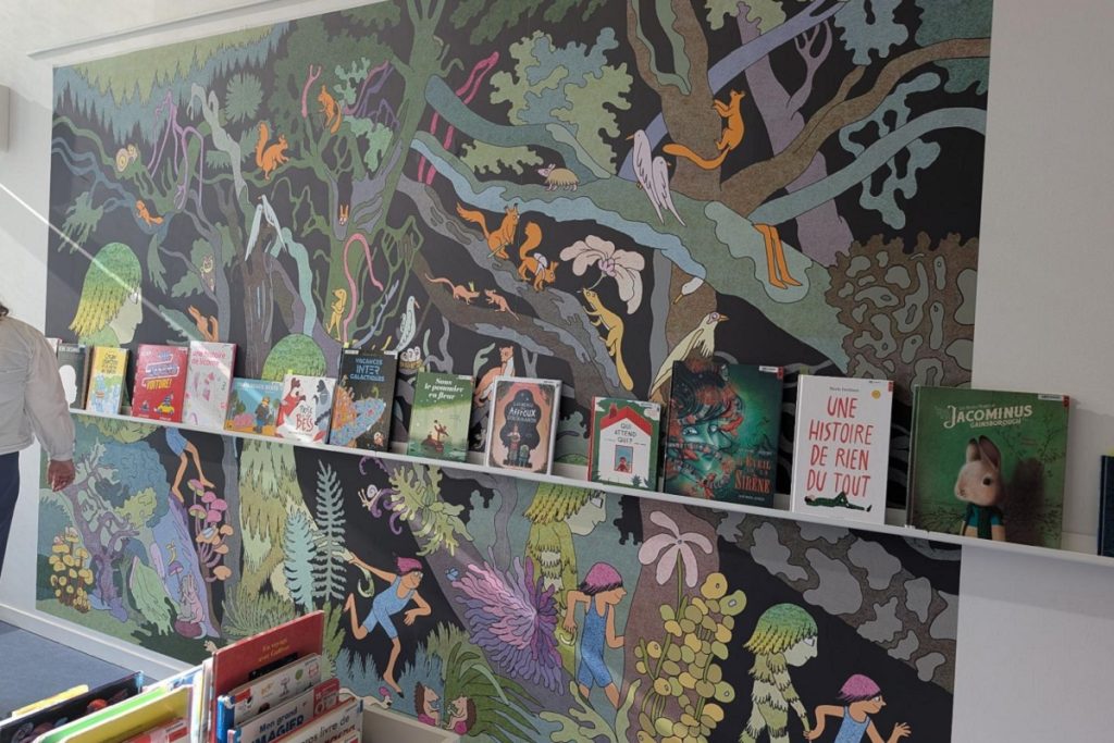 Fresque murale illustrée représentant une forêt peuplée d'animaux, surplombant une étagère de livres jeunesse dans l'espace jeunesse.