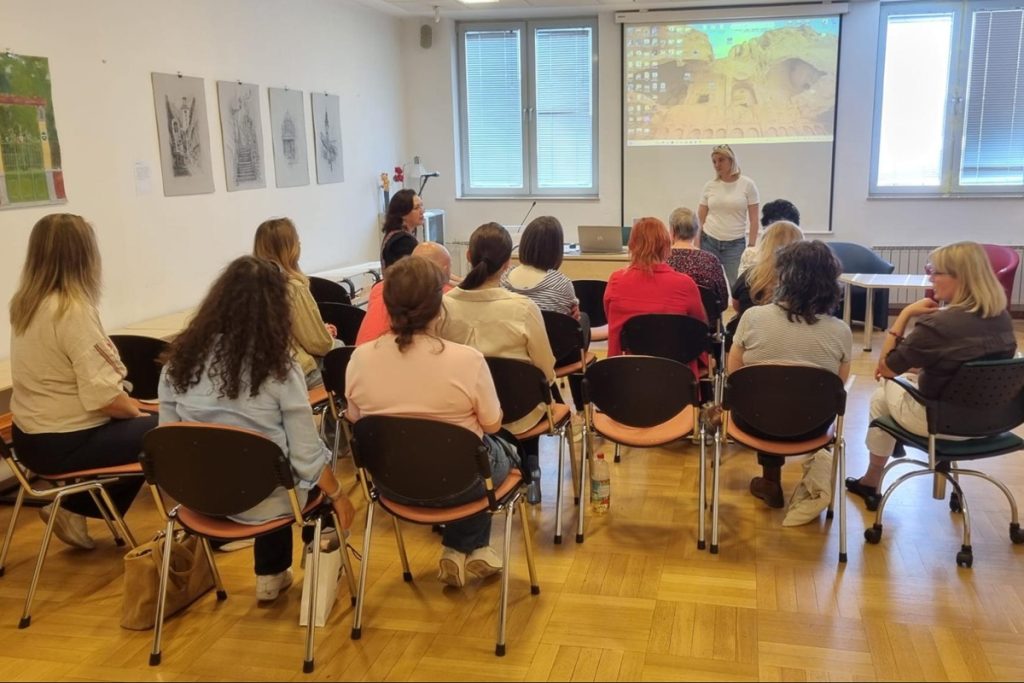 Salle dans la bibliothèque August Cesarec : une quinzaine de personnes assises en rangées de chaises écoutent une intervenante debout devant un écran de projection. Des dessins au crayon représentant des monuments sont accrochés au mur de gauche.