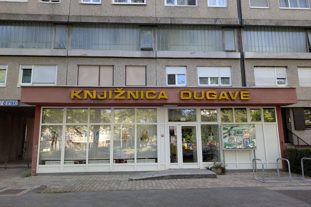 Façade de la bibliothèque de Dugave : un bâtiment résidentiel en béton gris des années soviétiques dont le rez-de-chaussée est occupé par la bibliothèque, signalée par une enseigne en lettres jaunes "Knjižnica Dugave". De larges vitrines donnent sur la rue pavée.