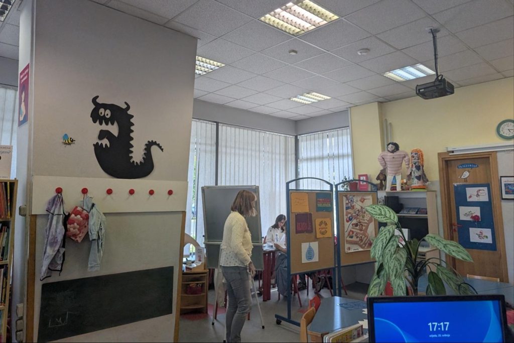 Salle dans la bibliothèque Marin Držić : une pièce lumineuse décorée d'un dragon noir en découpe murale. Deux femmes sont présentes, l'une debout près d'un tableau blanc, l'autre assise. Des panneaux d'affichage, des jouets et des plantes décorent l'espace. Un écran affiche 17h17.