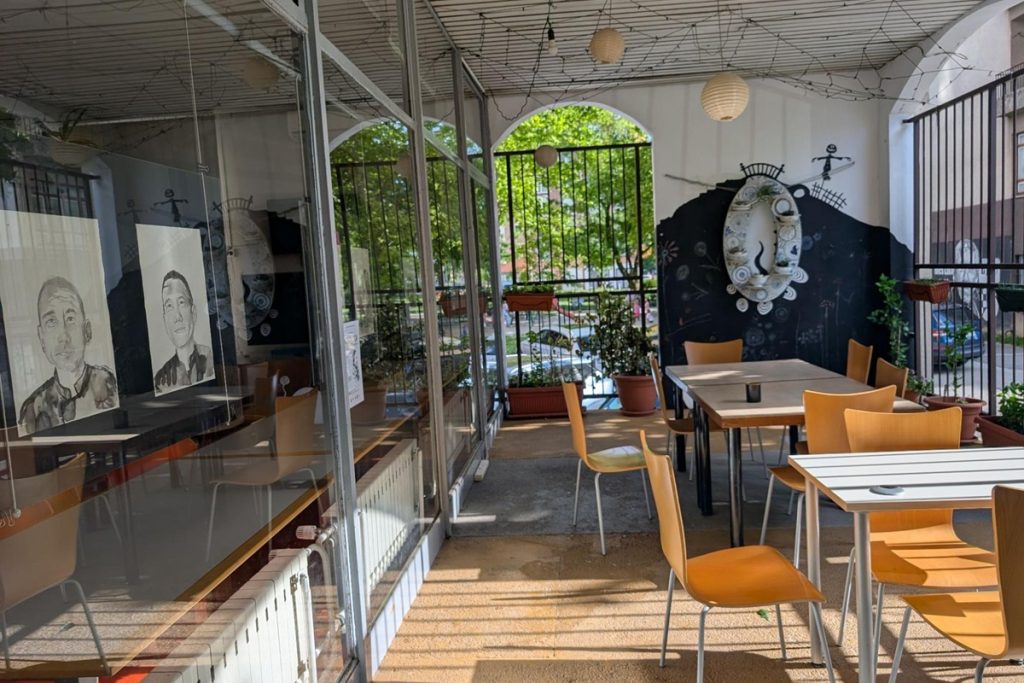 Espace semi-ouvert de la bibliothèque Voltino : des tables et chaises en bois clair sont disposées dans une véranda vitrée donnant sur un jardin verdoyant. Sur la paroi gauche, deux grands portraits dessinés au crayon de personnes migrantes. Un mur noir à droite est orné d'une œuvre en céramique.