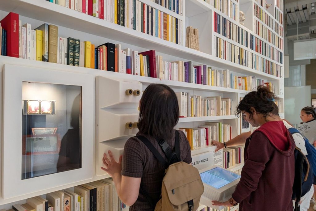 Bibliothèque murale avec visiteurs consultant les dispositifs manipulables installés parmi les rayonnages.