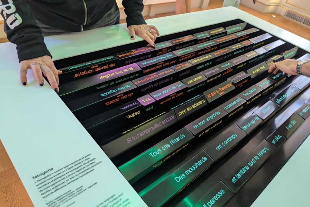 Table tactile lumineuse où deux visiteurs assemblent des bandeaux de mots colorés pour créer un texte à partir de plusieurs registres de langue.