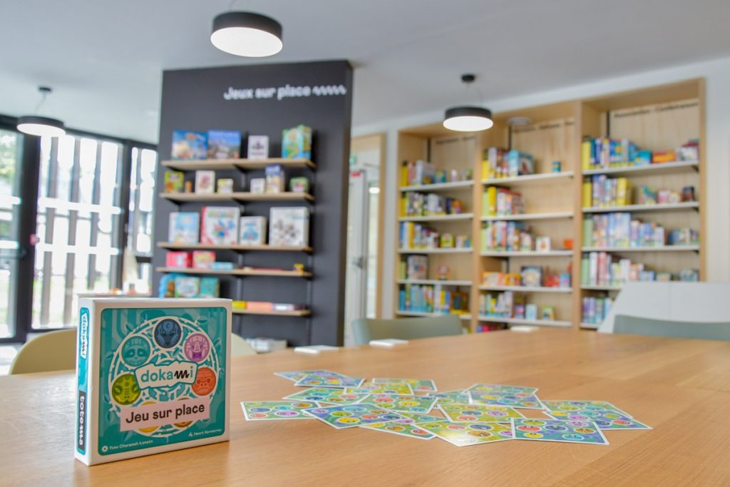 Jeu de société Dokami posé sur une table, avec en arrière-plan des étagères garnies de jeux à emprunter.