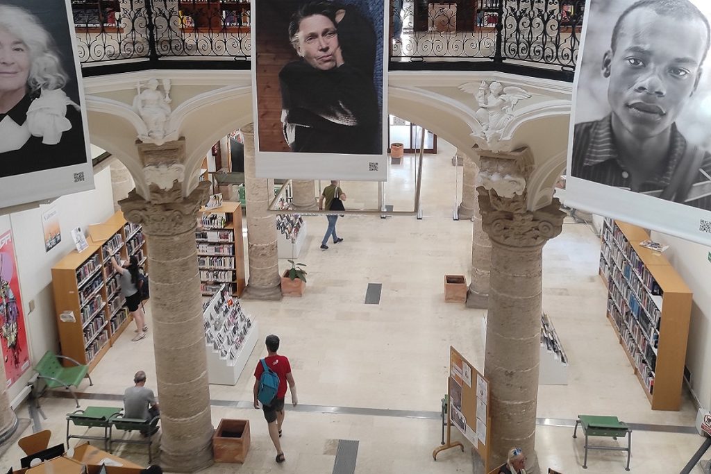 Vue plongeante sur le hall de la bibliothèque Pilar Faus, ancien hôpital du XVe siècle, avec colonnes en pierre et grands portraits suspendus.