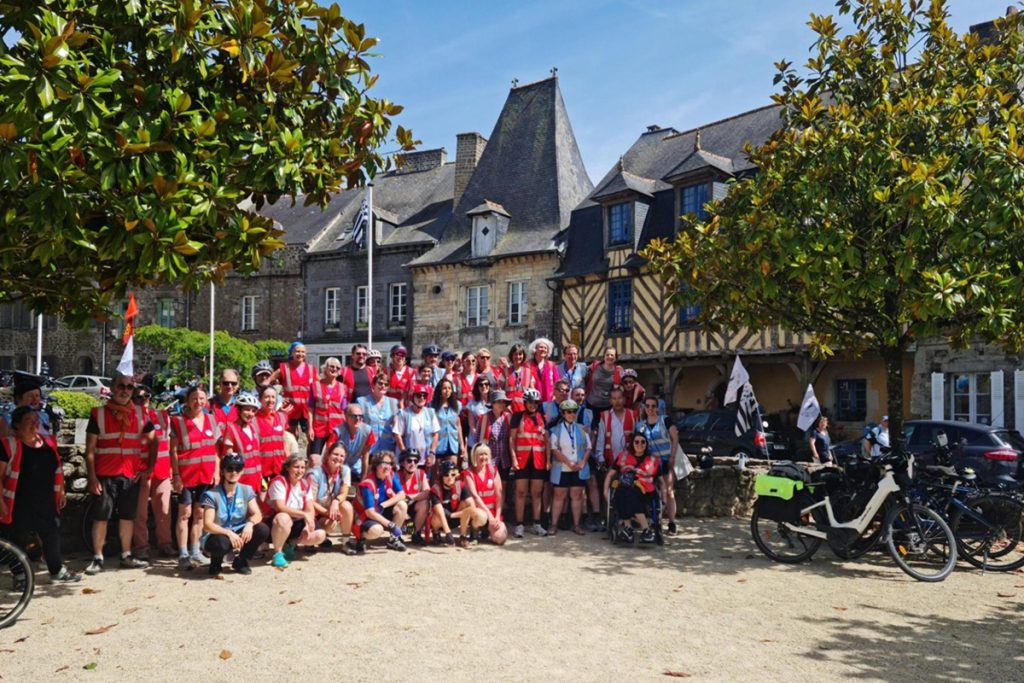 Groupe de bibliothécaires cyclistes en gilets rouges et bleus posant avec leurs vélos devant les maisons à pans de bois de Bécherel.