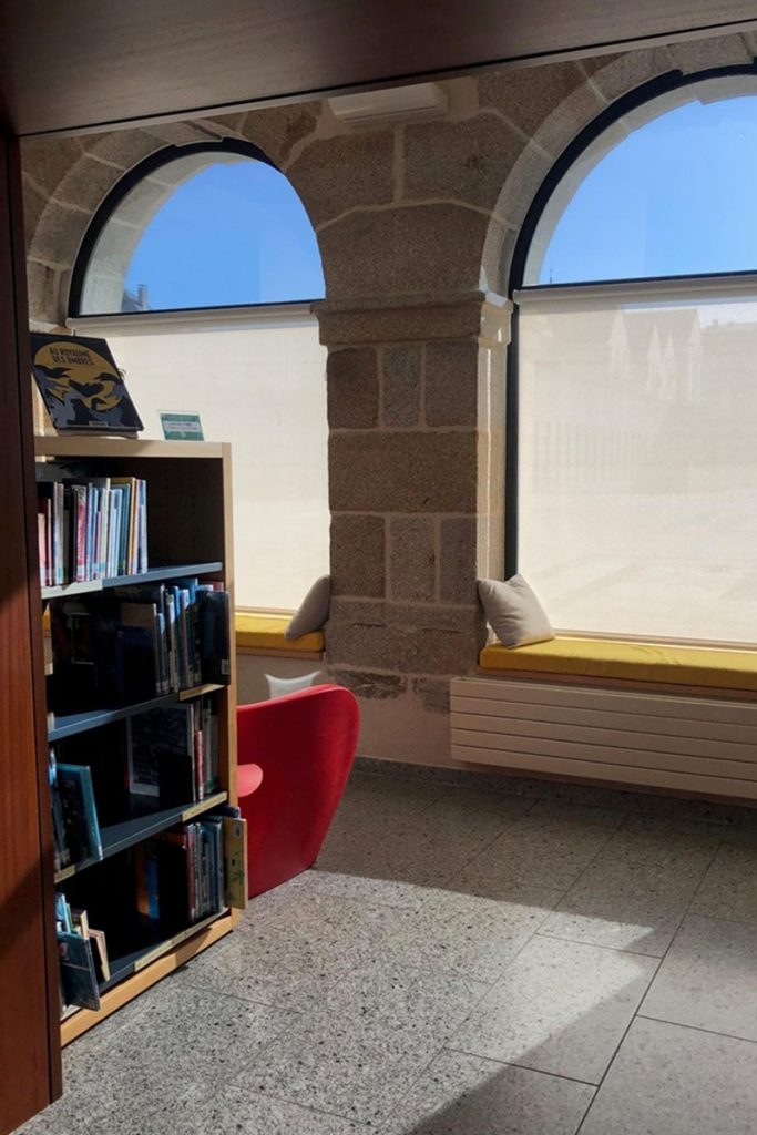 Coin lecture aménagé sous deux fenêtres en arcade de pierre, avec banquette coussinée, fauteuil rouge et étagère de bandes dessinées.