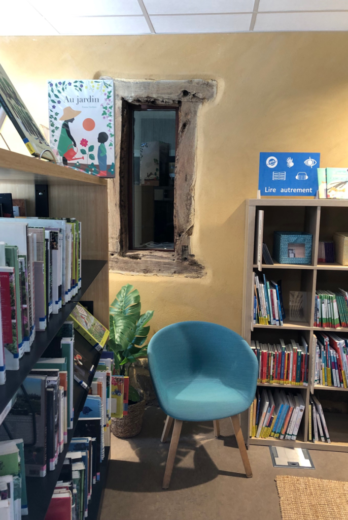 Coin lecture aménagé contre un mur ancien avec fenêtre en pierre et bois, fauteuil bleu et étagères de livres jeunesse.