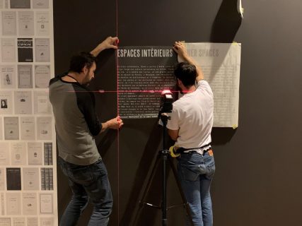 Photographie de deux personnes qui installent au mur un cartel d'exposition à l'aide d'un niveau laser