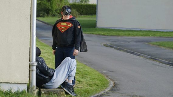 Un garçon se tient debout à l’angle d’une maison, avec un masque de Batman, une cape noire et un sweat Superman.