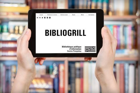 Deux mains portent une tablette devant une bibliothèque de livres sur laquelle est inscrit le nom Bibliogrill