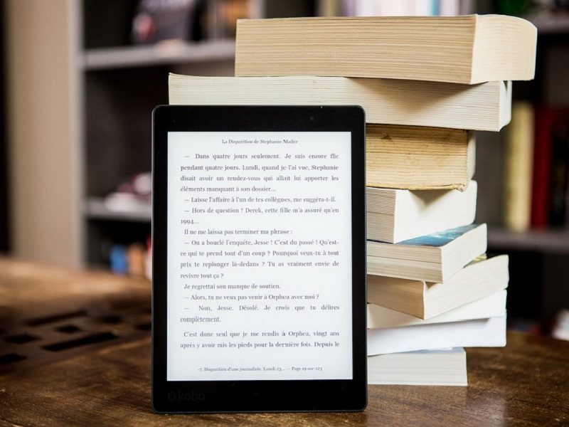 Une liseuse est posée verticalement devant une pile de livres papier.